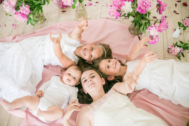 Cute children, a little brother and older sisters in white dresses lie together on the floor in a bright spring studio with pink peonies. Happy family. Happy childhood - Photo, image