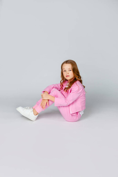 cute girl in a pink tracksuit and sneakers sits on a light gray background - Фото, изображение