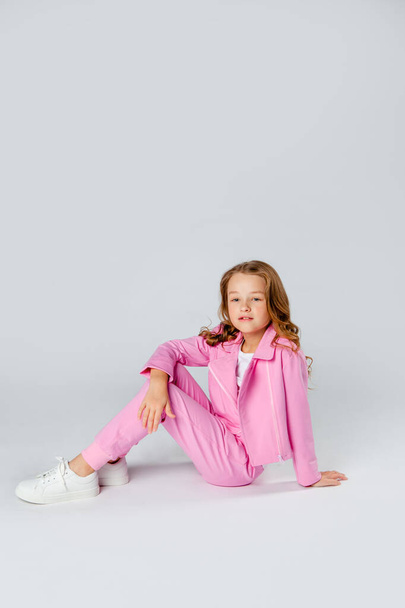 χαριτωμένο κορίτσι σε ένα ροζ φόρμα και αθλητικά παπούτσια κάθεται σε ένα ανοιχτό γκρι φόντο - Φωτογραφία, εικόνα