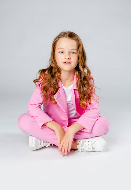 cute girl in a pink tracksuit and sneakers sits on a light gray background - Foto, Imagen