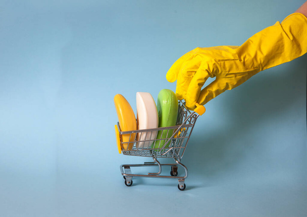 A female hand in a yellow glove rolls a supermarket trolley with pieces of soap on a blue background. Hygiene. Disinfection. Cleanliness. covid 19. - 写真・画像