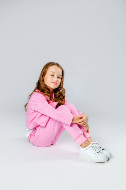 Pembe eşofmanlı ve spor ayakkabılı hoş bir kız açık gri bir arka planda oturuyor. - Fotoğraf, Görsel