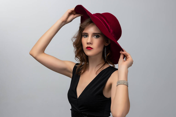 Beautiful photo of a young fashion model with long curly hair in a black long dress and red hat posing on grey background. Concept of hairstyling in salon and face care - Foto, imagen