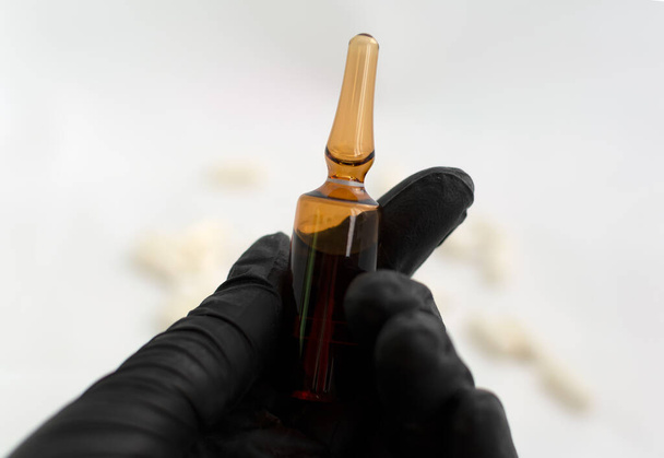 Hand in black latex glove holding medical glass ampule with medicine. Selective focus. Amber glass ampoule is held by hand in black medical glove with scattered pills at the background. Virus cure. - Photo, Image