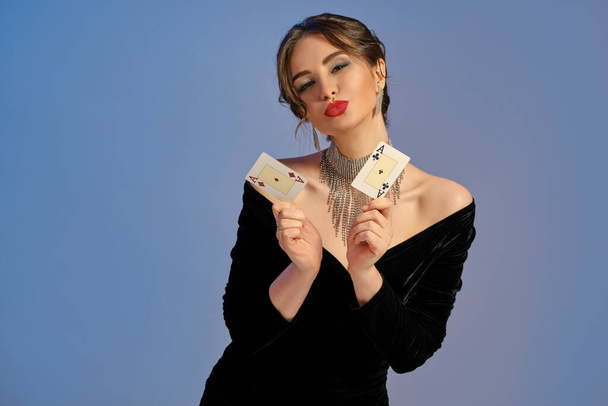 Alluring brunette lady with bare shoulders, bright make-up, in black dress and shiny jewelry. She is kissing you, showing two aces, posing against blue background. Poker, casino. Close-up, copy space - 写真・画像