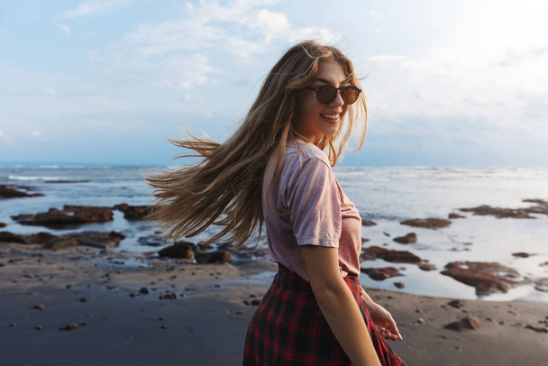 Romantikus vonzó, mosolygós hipster lány utazó, hátizsák élvezi a nyári vakáció, forduljon vissza, hogy néz vissza a vigyorral, séta a fekete homokos vulkanikus strand Indonézia trópusi üdülőhely - Fotó, kép
