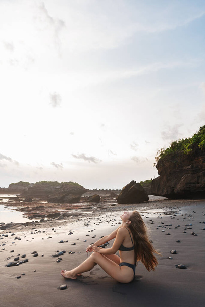 Tropical island, vacation and summer lifestyle concept. Vertical shot of sexy young carefree woman in swimsuit, sit on black volcanic sand at beach, enjoying blue ocean landscape, waves, sunbathing - 写真・画像