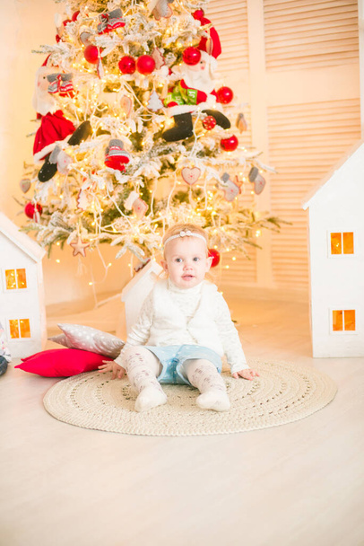 Χαριτωμένο κοριτσάκι με ξανθά μαλλιά παίζει σε ένα φωτεινό δωμάτιο διακοσμημένο με χριστουγεννιάτικες γιρλάντες κοντά στο χριστουγεννιάτικο δέντρο. Ευτυχισμένη παιδική ηλικία. Χριστουγεννιάτικη διάθεση - Φωτογραφία, εικόνα