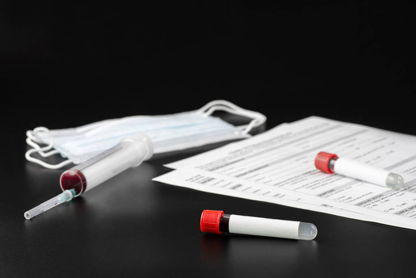 Two vacuum blood test tubes with a blank label for inserting text, a syringe and a protective face mask on a black background. The concept covid-19 or the concept of coronavirus - Photo, Image
