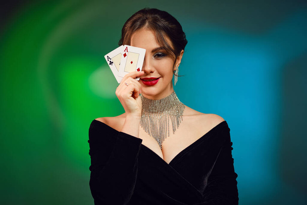 Brunette girl with bare shoulders and bright make-up. In black dress, brilliant necklace and earrings. Smiling, covered her eye by two aces, posing on colorful background. Poker, casino. Close-up - Фото, зображення