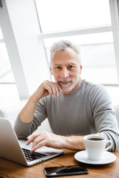 Afbeelding van een knappe positieve volwassen senior zakenman zitten in cafe met behulp van laptop computer. - Foto, afbeelding