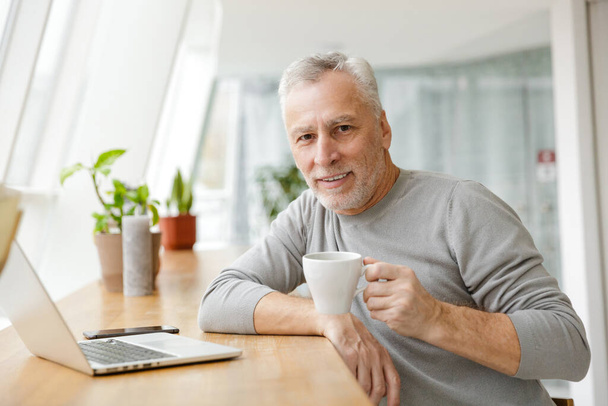 Image of a positive mature senior businessman sit in cafe using laptop computer drinking coffee. - Fotó, kép