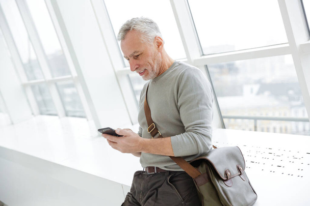 Foto van een senior optimistische grijs-harige bebaarde zakenman binnen chatten met een mobiele telefoon. - Foto, afbeelding