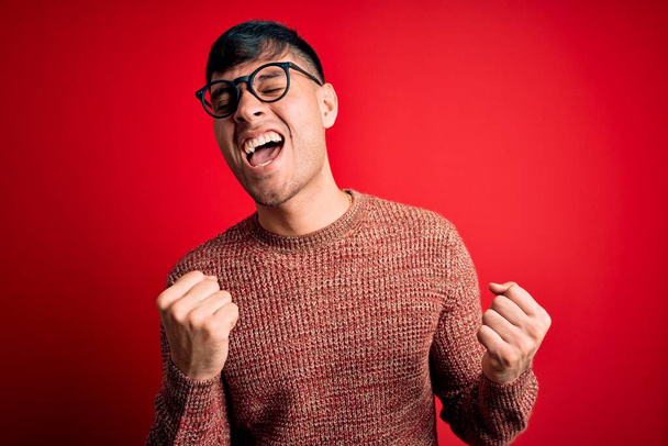 Homem hispânico bonito jovem usando óculos nerd sobre fundo vermelho muito feliz e animado fazendo gesto vencedor com os braços levantados, sorrindo e gritando para o sucesso. Conceito de celebração
. - Foto, Imagem