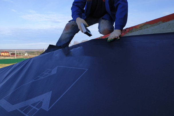 De arbeider wikkelt een rol isolatiefilm af en bedekt deze met een dak.2020 - Foto, afbeelding