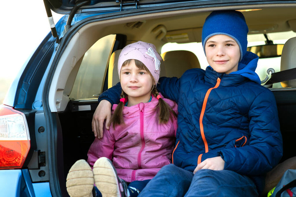 Два счастливых мальчика и девочка сидят вместе в багажнике машины. Веселые брат и сестра обнимаются в семейном багажном отделении. Путешествия на выходные и праздники. - Фото, изображение