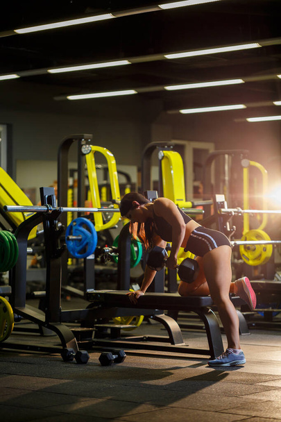 The girl in a black sports suit performs an exercise with dumbbells in the gym - Foto, imagen