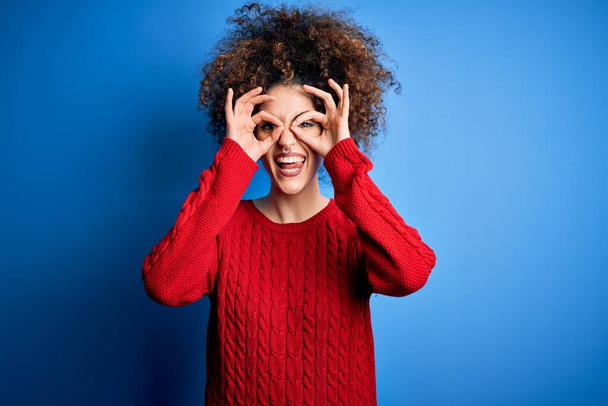 Young beautiful woman with curly hair and piercing wearing casual red sweater doing ok gesture like binoculars sticking tongue out, eyes looking through fingers. Crazy expression. - Zdjęcie, obraz