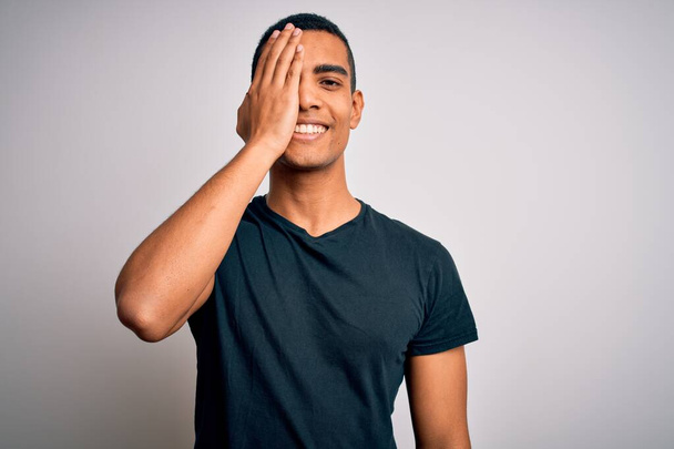 Jovem homem afro-americano bonito vestindo camiseta casual em pé sobre fundo branco cobrindo um olho com a mão, sorriso confiante no rosto e emoção surpresa
. - Foto, Imagem