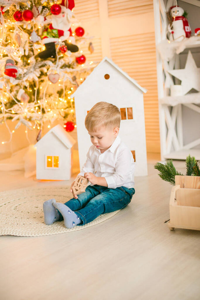 Cute little boy with blond hair plays with  toys in a bright room decorated with Christmas garlands near the Christmas tree. Happy childhood. Christmas mood - Фото, зображення