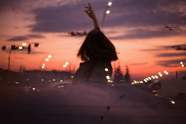 Művészeti fotó. Esti fotó egy lányról az út mellett. A lány örvénylik és élvezi az életet. Fények égnek, autók vezetnek.Naplemente, az ég világos és telített színek.Reflection autók. A lány haja fejlődik. - Fotó, kép