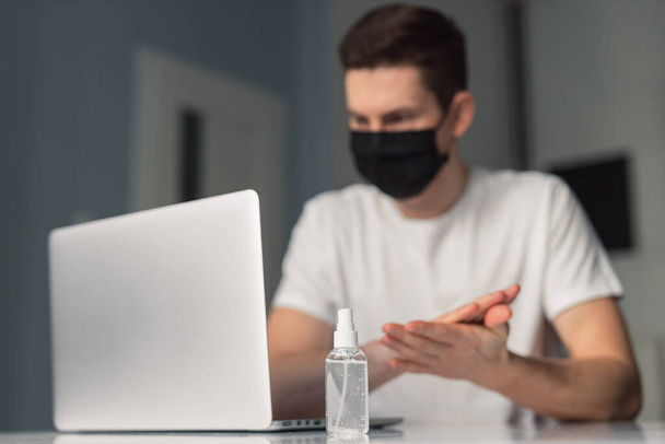 Man working from home during coronavirus quarantine, wearing black protective mask. Working from home. Cleaning her hands with sanitizer gel. Close up photo of sanitizer gel - Fotografie, Obrázek