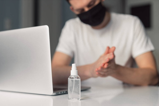 Man working from home during coronavirus quarantine, wearing black protective mask. Working from home. Cleaning her hands with sanitizer gel. Close up photo of sanitizer gel - Photo, Image
