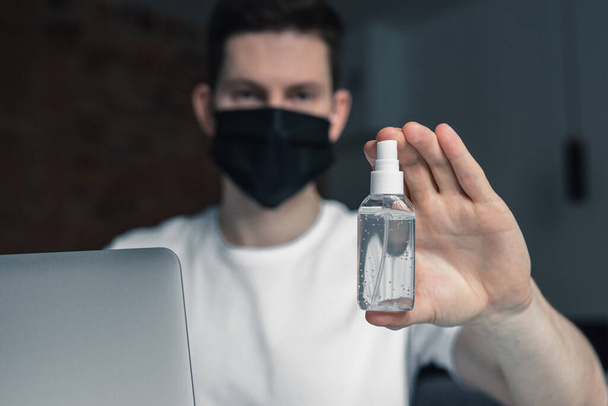 Man working from home during coronavirus quarantine, wearing black protective mask. Working from home. Cleaning her hands with sanitizer gel. Close up photo of sanitizer gel - Photo, image