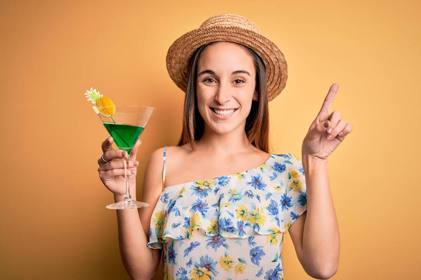 Joven hermosa turista de vacaciones con sombrero de verano beber bebida cóctel sorprendido con una idea o pregunta dedo con la cara feliz, número uno
 - Foto, Imagen