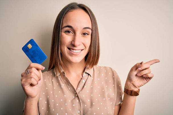 Junge schöne Kundin, die Kreditkarte hält, um auf weißem Hintergrund zu bezahlen, zeigt sehr glücklich mit Hand und Finger zur Seite - Foto, Bild