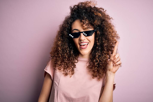 Young beautiful woman with curly hair and piercing wearing funny thug life sunglasses pointing finger up with successful idea. Exited and happy. Number one. - Foto, Imagem