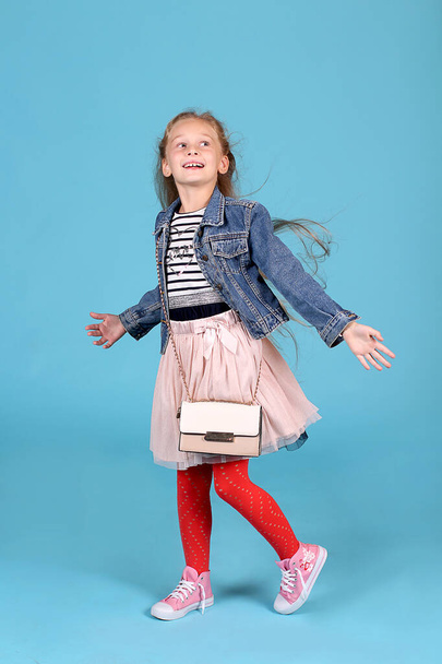 Μικρή fashionista με μια τσάντα θέτει σε ένα μπλε φόντο. Ριγέ T-shirt με denim σακάκι, φούστα, κόκκινο καλσόν και ροζ sneakers. Καρφιτσώστε. - Φωτογραφία, εικόνα