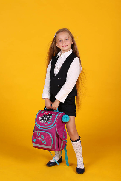 Portrait of a first-grader girl in a school uniform with long flowing hair on a yellow background with a backpack - Photo, Image