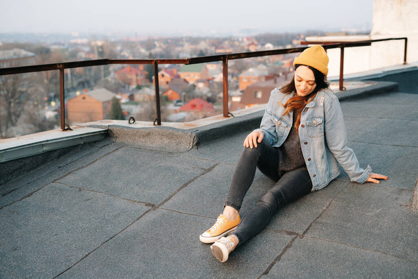 pretty white European woman is sitting on the roof of a multi-storey building in a blue denim jacket in gray pants,a yellow stylish hat.in the evening at sunset. lifestyle concept. trendy seasons - Photo, Image