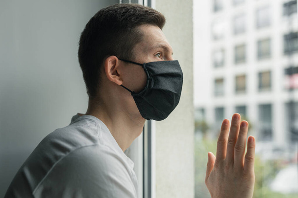 Man looking from window during coronavirus quarantine, wearing black protective mask. Working from home.  - Foto, imagen