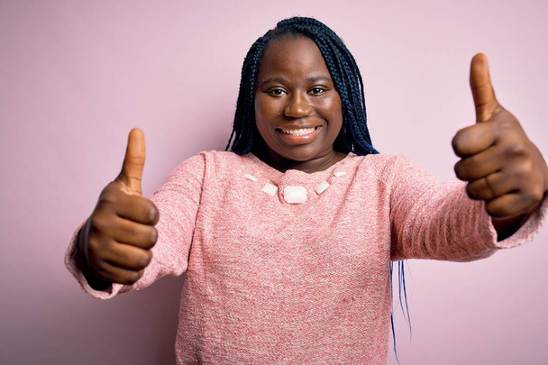 Африканский американец плюс размер женщины с косичками носить случайный свитер на розовом фоне одобряя делать положительный жест с рукой, пальцы вверх улыбается и счастлив за успех. Жест победителя
. - Фото, изображение