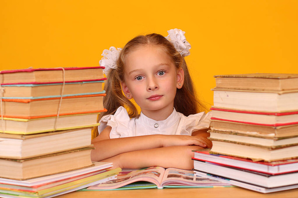 Birinci sınıf öğrencisi bir kızın portresi sarı arka plandaki kitapların arasında bir masada oturuyor. - Fotoğraf, Görsel