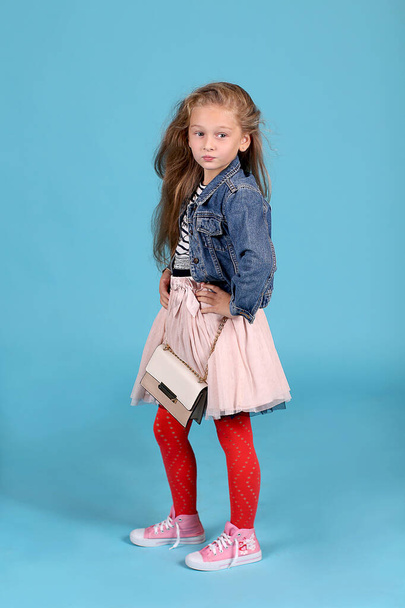 Little fashionista with a handbag posing on a blue background. Striped T-shirt with denim jacket, skirt, red tights and pink sneakers. Pin up. - 写真・画像