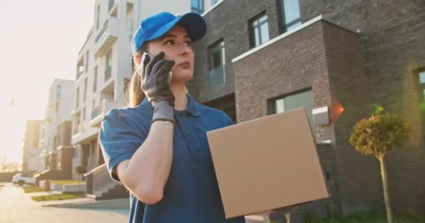 Caucasian young pretty woman, delivery worker in blue hat cap walking the street, carrying cardbox parcel and talking on mobile phone. Female beautiful courier with carton box speaking on cellphone. - Video