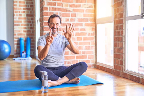 Красивый спортсмен средних лет сидит на коврике и делает упражнения йоги на растяжку в спортзале, показывая и указывая пальцами номер шесть, улыбаясь уверенно и счастливо
. - Фото, изображение