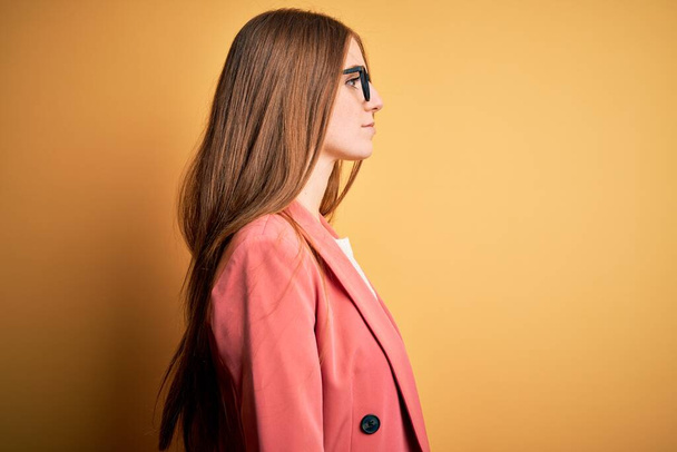 Fiatal gyönyörű vörös hajú nő visel kabát és szemüveg felett elszigetelt sárga háttér keres oldalra, pihenjen profil pózol természetes arc magabiztos mosollyal. - Fotó, kép