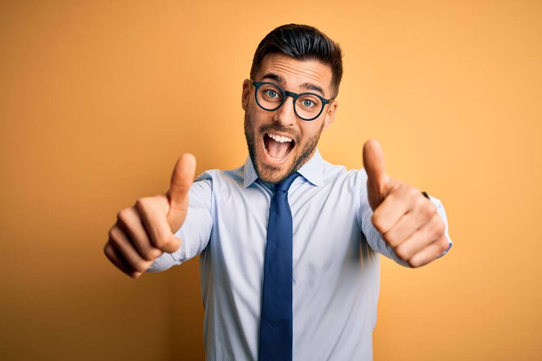 Junge gutaussehende Geschäftsleute mit Krawatte und Brille stehen vor gelbem Hintergrund und genehmigen positive Gesten mit der Hand, Daumen hoch lächelnd und glücklich über den Erfolg. Siegergeste. - Foto, Bild