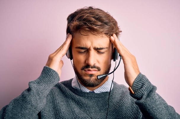 Mladý agent call centra muž s vousy nosí sluchátka přes izolované růžové pozadí trpí bolestí hlavy zoufalý a ve stresu, protože bolest a migréna. Ruce za hlavu. - Fotografie, Obrázek