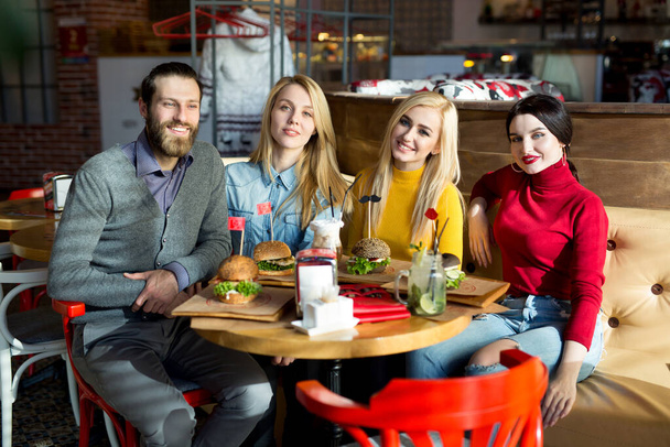 Οι άνθρωποι τρώνε μαζί σ 'ένα τραπέζι σ' ένα καφέ. Ευτυχισμένοι φίλοι τρώνε χάμπουργκερ και πίνουν κοκτέιλ στο εστιατόριο - Φωτογραφία, εικόνα