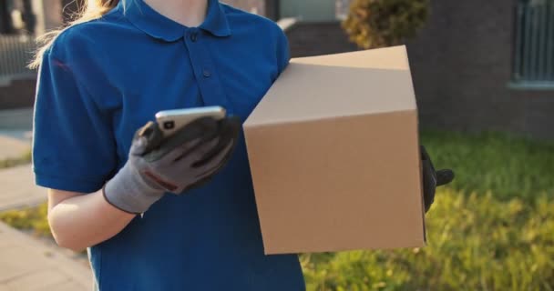 Caucasian young pretty woman, delivery worker in cap walking the street, carrying carton box and tapping on smartphone. Female beautiful courier with parcel texting or scrolling on phone. Outdoor. - Video