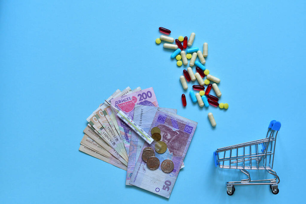 Shopping trolley cart with assorted medicine pills,medical injection and Ukrainian hryvnia on blue background. Creative idea for drugstore, online pharmacy. Flat layout.Selective Focus.Coronavirus - Foto, Bild