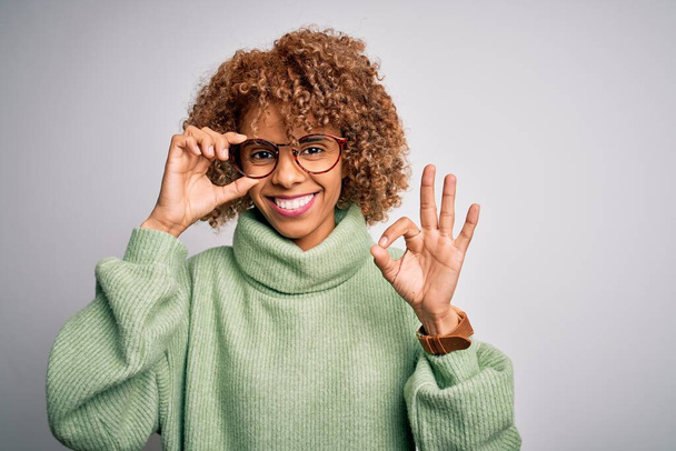 Africká americká optická žena s kudrnatými vlasy nosí brýle na izolovaném bílém pozadí dělá v pořádku znamení s prsty, vynikající symbol - Fotografie, Obrázek