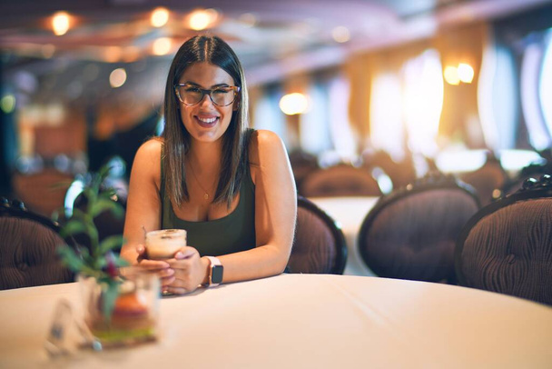 Молодая красивая женщина улыбается счастливой и уверенной. Сидя с улыбкой на лице держа стакан кофе в ресторане
 - Фото, изображение