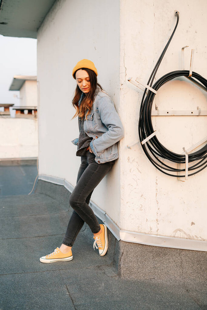 white European woman stands near the wall on the roof of a multi-storey building in stylish denim clothing and a trendy yellow hat .in the rays of the setting sun.lifestyle concept. fashion seasons - Фото, изображение