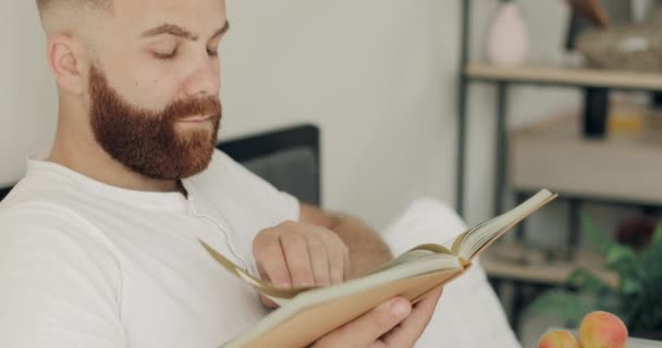 Close up view of concentrated young man reading novel while having breakfast in bed. Handsome bearded guy in middle 30s sitting and turning book page. Concept of leisure. - Imágenes, Vídeo